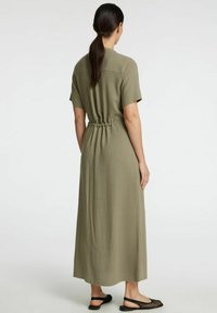 Robe maxi vert olive avec des manches courtes, une taille froncée et une silhouette droite, fabriquée en tissu léger. Chaussures plates noires.
