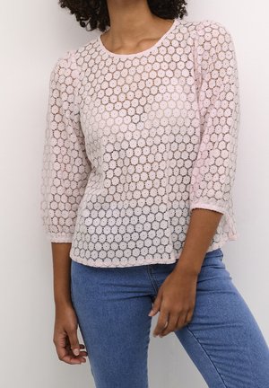 Femme portant un haut en dentelle rose clair avec des motifs circulaires et des manches trois-quarts, associé à un jean bleu.