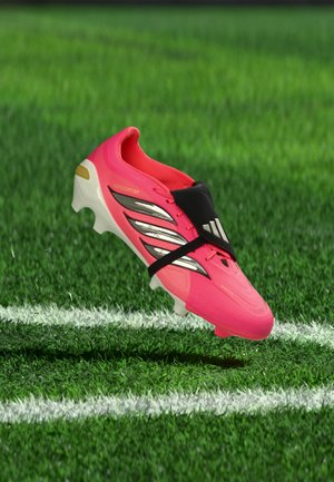 Crampon de football rose vif avec des rayures noires et blanches flottant au-dessus de l'herbe verte près d'une ligne de touche blanche.