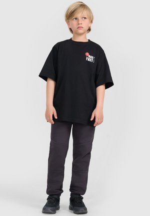 Blond dziecko stoi, nosi czarny oversize t-shirt z napisem "basket" oraz ciemnoszare spodnie i czarne tenisówki, patrzy na bok.