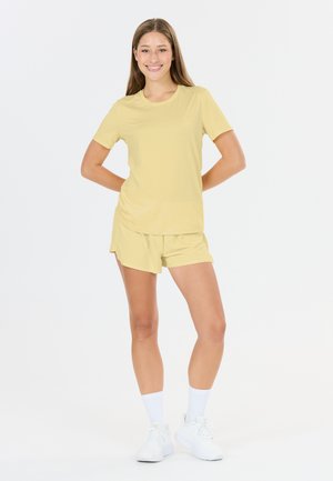 Junge Frau steht, lächelt, trägt ein hellgelbes T-Shirt und passende Shorts mit weißen Socken und Turnschuhen auf weißem Hintergrund.