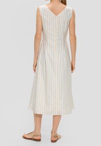 Mujer con un vestido midi sin mangas de rayas verticales en beige y blanco, combinado con sandalias planas doradas, de pie frente a un fondo liso.