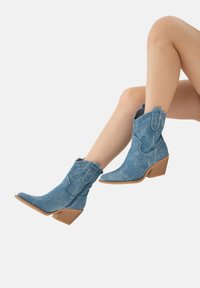 Botas de cowboy de ante azul claro con puntas afiladas, tacones angulados y costuras decorativas. Presentan cremalleras laterales y un acabado ligeramente desgastado.