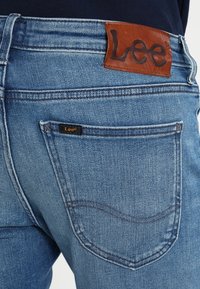 Denim jeans in light blue, featuring a brown leather patch with "Lee" logo, back pocket with stitching detail, and standard fit design.