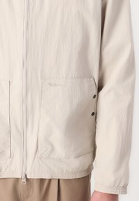 Beige lichtgewicht jack met een gestructureerde afwerking, rits aan de voorkant, twee grote zakken en elastische zoom en manchetten. Bevat een subtiel merkembleem.