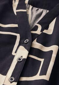 Kurzarmhemd aus schwarzem Stoff mit cremefarbenem, abstraktem geometrischen Muster. Verfügt über eine Knopfleiste und eine weiche Textur.