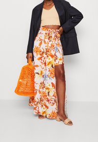 Žena v černém blejzru, béžovém cropped topu, květinové sukni s rozparkem na stehně, béžových sandálech na podpatku a držící jasně oranžovou tkanou tašku.