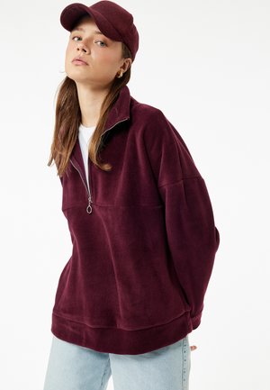 Burgundowy polarowy sweter z kołnierzem na suwak do połowy, opadającymi ramionami, miękką fakturą oraz luźnym krojem, w zestawieniu z pasującą czapką.