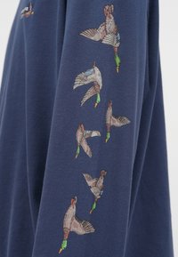 Tumed sinine dress, millel on detailne brodeeritud kujutis lendavatest partest varrukal, esitleb erinevaid värve ja tekstuure.