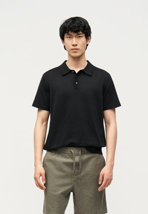 Fiatal férfi fekete rövid ujjú galléros ingben és olívazöld zsinóros rövidnadrágban, világos egyszínű háttér előtt áll.