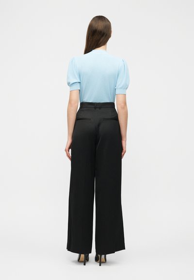 Μια γυναίκα στέκεται γυρισμένη από την πλάτη, φορώντας ένα ανοιχτό μπλε τοπ με φουσκωτά μανίκια, μαύρο παντελόνι με φαρδιά μπατζάκια και μαύρα ψηλοτάκουνα παπούτσια.