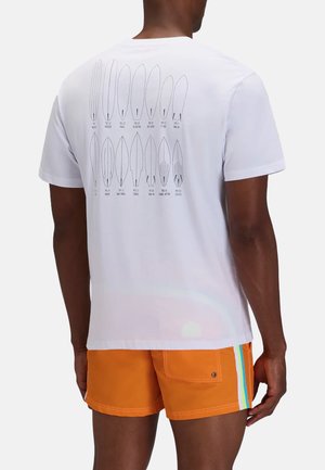 Uomo che indossa una maglietta bianca con illustrazioni tipo tavola da surf e pantaloncini arancioni con striscia blu e bianca sul lato, visto da dietro.
