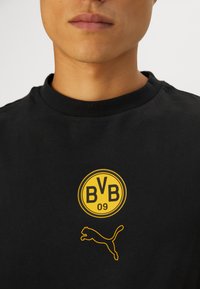 Sort bomulds t-shirt med et gult cirkulært logo med "BVB 09" og en gul puma-silhuet i nederste højre hjørne. Rund halsudskæring.