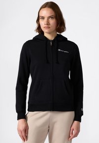 Sudadera con cremallera negra, hecha de un material suave, con un bolsillo en la parte frontal, capucha con cordón y un logo bordado en blanco en el pecho y la manga.