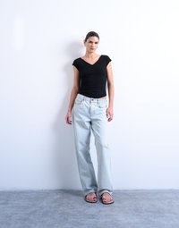 Γυναίκα που στέκεται μπροστά σε έναν λευκό τοίχο φορώντας μαύρο μπλουζάκι με V λαιμόκοψη, ανοιχτόχρωμο τζιν και σανδάλια πάνω σε γκρι δάπεδο.