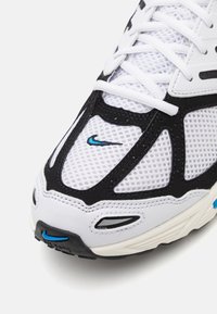 Bílé a černé sportovní boty s síťovinou, bílé tkaničky a malé modré logo Nike swoosh na špičce a přední části podrážky.