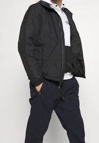 Černá péřová bunda má zip vpředu, boční kapsy a elastické manžety. Je nošena přes světle šedou mikinu a doplněna námořnickými kalhotami.