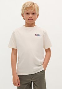 Lichtcrème t-shirt van zacht katoen met korte mouwen, voorzien van een klein paars en blauw logo op de linkerkant van de borst. Gemaakt met groene broek.