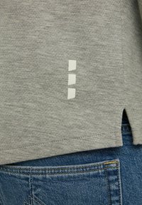 Graues kurzärmliges Hemd mit strukturierter Oberflächenbeschaffenheit, das drei weiße quadratische Akzente an der Seite und einen kleinen seitlichen Schlitz aufweist. Denim ist darunter sichtbar.