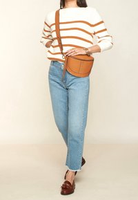 Tasche aus tannfarbigem Leder mit Nietenakzenten, cremefarbener Pullover mit orangefarbenen Streifen, hellblaue gerade Jeans und braune Mokassins.