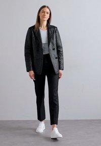 Giacca nera trapuntata doppiopetto con una texture a motivo; abbinata a pantaloni neri e scarpe bianche; maglia grigia chiara sotto.