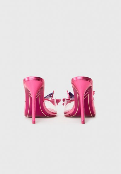 Ein Paar pinkfarbene High Heels Sandalen mit klaren Riemen und Schmetterlingsdekorationen, von hinten auf einer weißen Oberfläche betrachtet.