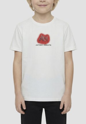 Bambino che indossa una t-shirt bianca con logo Antony Morato rosso e nero, abbinata a pantaloni neri, in piedi davanti a uno sfondo chiaro uniforme.