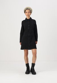 Crna traper haljina sa ovratnikom, prednjim kopčanjem na dugmad, dugim rukavima i uskim strukom, koja završava iznad koljena. Upari se s crnim čizmama s vezicama.