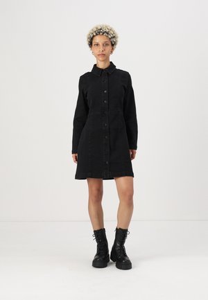 Svart denimkjole med krage, frontlukking med knapper, lange ermer og figursydd midje, som slutter over kneet. Kombinert med svarte snøresko.