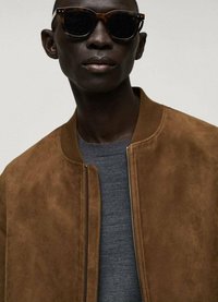 Giacca bomber in suede marrone con chiusura a zip, abbinata a un maglione grigio. Modello che indossa occhiali da sole tartarugati, evidenziando una superficie liscia.