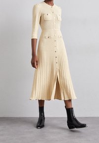 Krämfärgad stickad klänning med texturerat mönster, knäppning med knappar, två bröstfickor och plisserad kjol. Buren med svarta ankelstövlar.