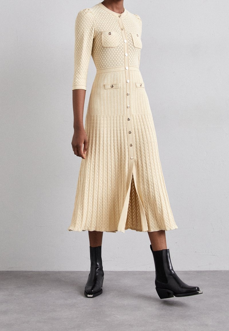 Krämfärgad stickad klänning med texturerat mönster, knäppning med knappar, två bröstfickor och plisserad kjol. Buren med svarta ankelstövlar.