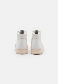 Un par de zapatillas deportivas altas blancas con suelas beige vistas desde atrás sobre un fondo blanco liso.