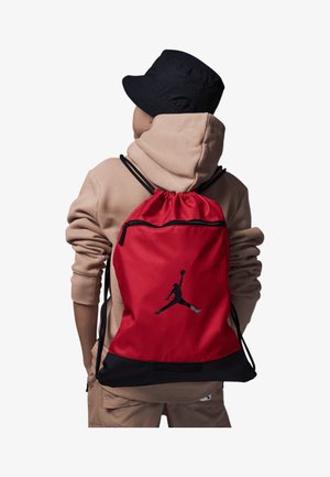 Sac à dos rouge à cordon de serrage avec des accents noirs, présentant une silhouette noire d'une figure sautant. Fabriqué en matériau durable avec une sangle ajustable.