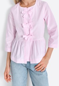 Světle růžová blůza s bílým pruhováním, třemi mašlemi, tříčtvrtečními rukávy, zaobleným výstřihem a peplum designem.