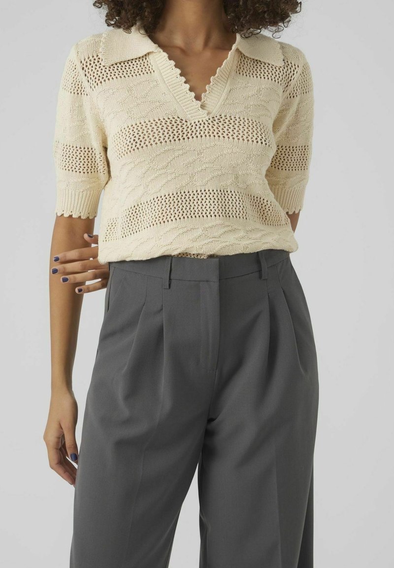 Femme portant un haut en maille crème à manches courtes et col en V rentré dans un pantalon plissé gris taille haute.