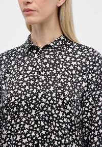 Zwart-witte blouse met een ster- en ovale patroon, voorzien van een kraag en knopen aan de voorkant. Gladde stoftextuur.