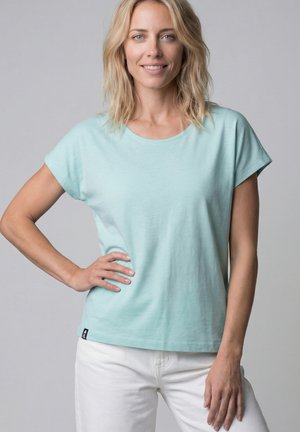 Blonde Frau in einem hellblauen kurzärmligen Hemd und weißen Hosen steht mit einer Hand in der Hüfte und lächelt in die Kamera vor einfachem Hintergrund.