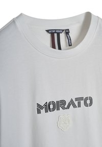 Camiseta de algodón blanca con cuello redondo, que presenta el texto negro "MORATO" y un diseño bordado en blanco que se asemeja a una flor o un animal.