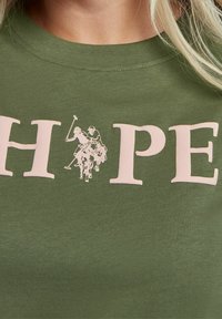 Close-up of green T-shirt with "HOPE" text and two polo players on horses printed between the letters.
