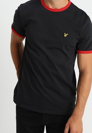 Mand iført sort t-shirt med rød krave og ærmekanter, med en lille gul fuglelogo på venstre bryst.