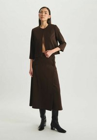 Veste courte marron à manches trois quarts, associée à une jupe marron taille haute avec des boutons, complétée par des bottes noires jusqu'aux genoux.