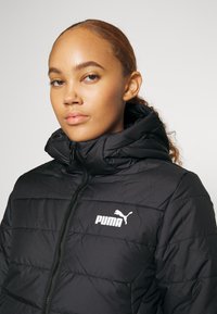 Mustmust jakk, millel on kõrge kaelus, tikandiga disain ja valge Puma logo rinnal. Ees asuv tõmblukk ja kapuuts lisavad soojust.