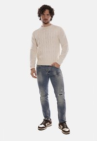 Maglione a maglia crema con motivo a trecce, abbinato a jeans azzurri chiari strappati e scarpe da ginnastica marroni e bianche.