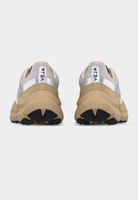 Ζευγάρι μπεζ και γκρι chunky αθλητικά παπούτσια με το λογότυπο «VEJA» στις ταμπέλες της φτέρνας, απεικονιζόμενα από πίσω σε λευκό φόντο.