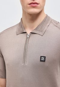 Κοντινό πλάνο ενός ατόμου που φοράει κοντομάνικη πόλο μπλούζα σε χρώμα ταουπέ με φερμουάρ και μαύρο τετράγωνο λογότυπο «HUGO» στο στήθος.