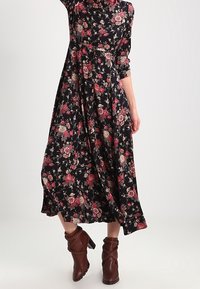 Donna che indossa un lungo abito floreale nero con fiori rossi e beige abbinato a stivali ankle con tacco marroni, in piedi contro uno sfondo semplice.