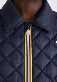 Giacca trapuntata blu navy con un colletto prominente, caratterizzata da una zip con dettaglio a strisce multicolori e hardware argentato. Materiale con texture visibile.