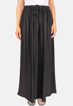Femme portant un pantalon large noir ample avec taille élastique et cordon, associé à des sandales noires à bout ouvert et un haut blanc.