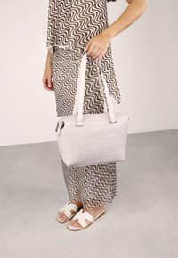 Bolso de mano rosa claro con un acabado texturizado, forma rectangular y dos correas para el hombro, sostenido por una persona que lleva un atuendo estampado y sandalias.